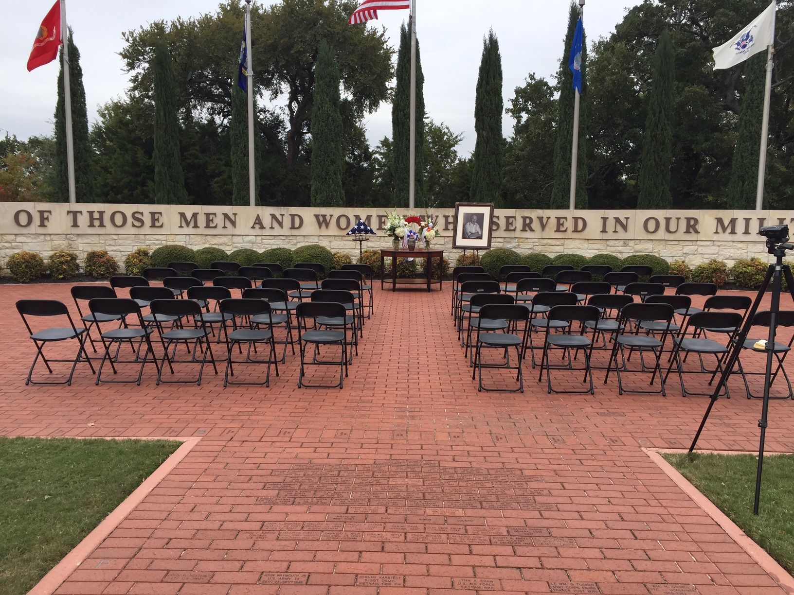Rows of black chairs face a memorial with flags. A small table holds items, a brick plaza setting.