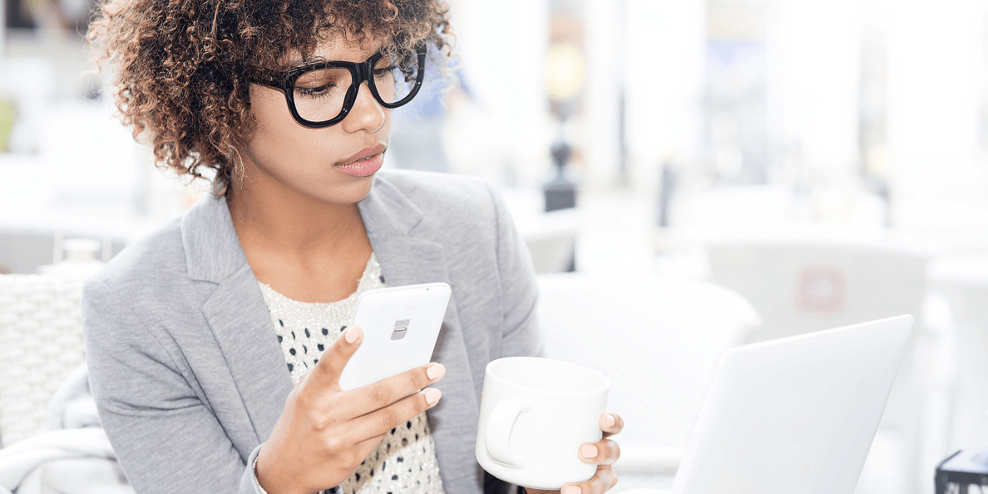 Person wearing glasses and a gray cardigan using a smartphone while holding a white mug in a bright office