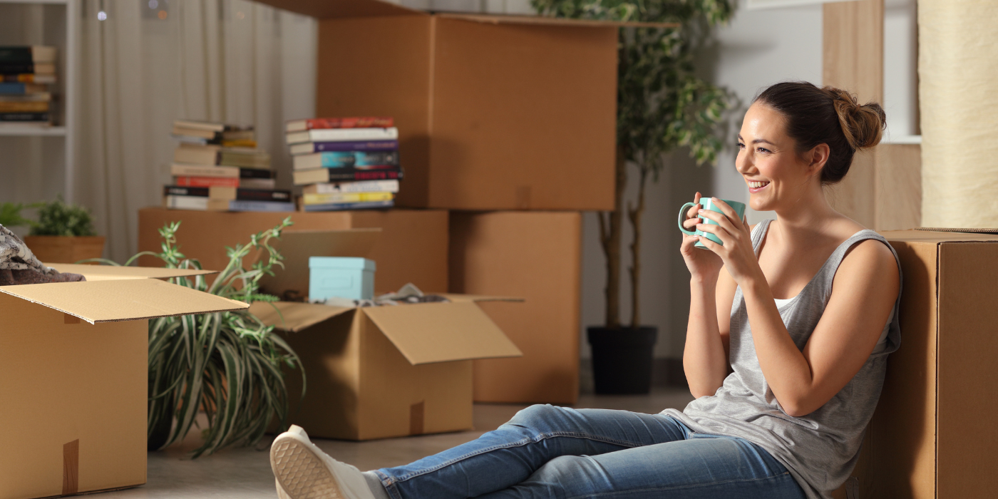 Woman sitting on a couch, smiling and holding a mug among packed moving boxes in a bright room