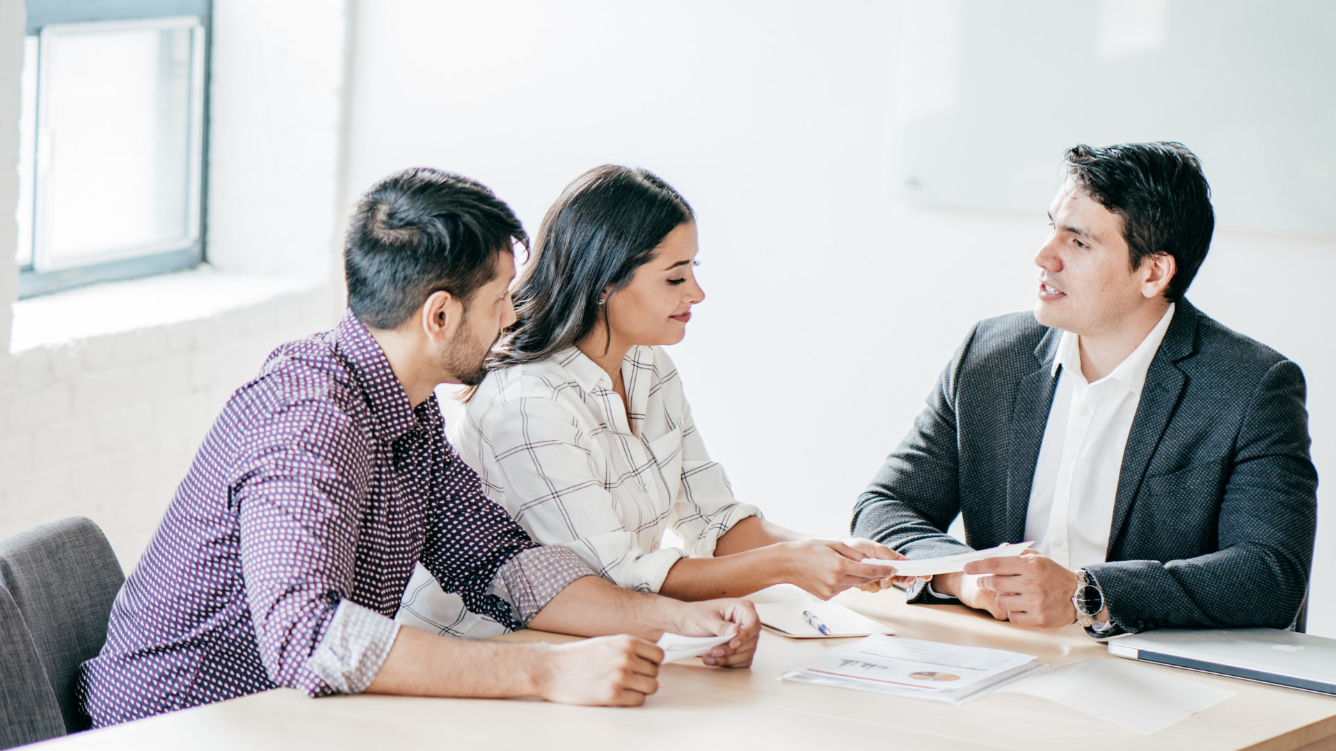 Three colleagues in a bright office meeting, leaning over papers and discussing a document.