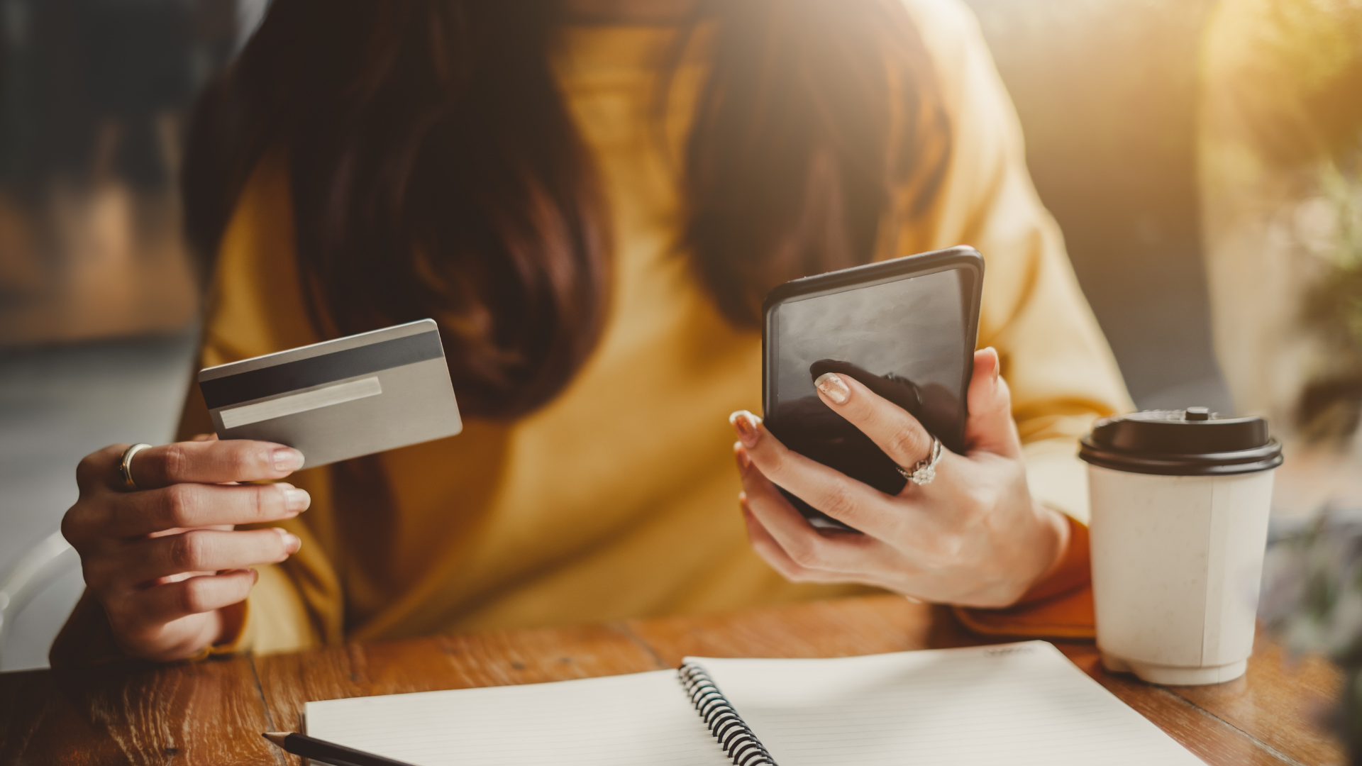 Person holding a credit card and smartphone at a table with coffee and a notebook