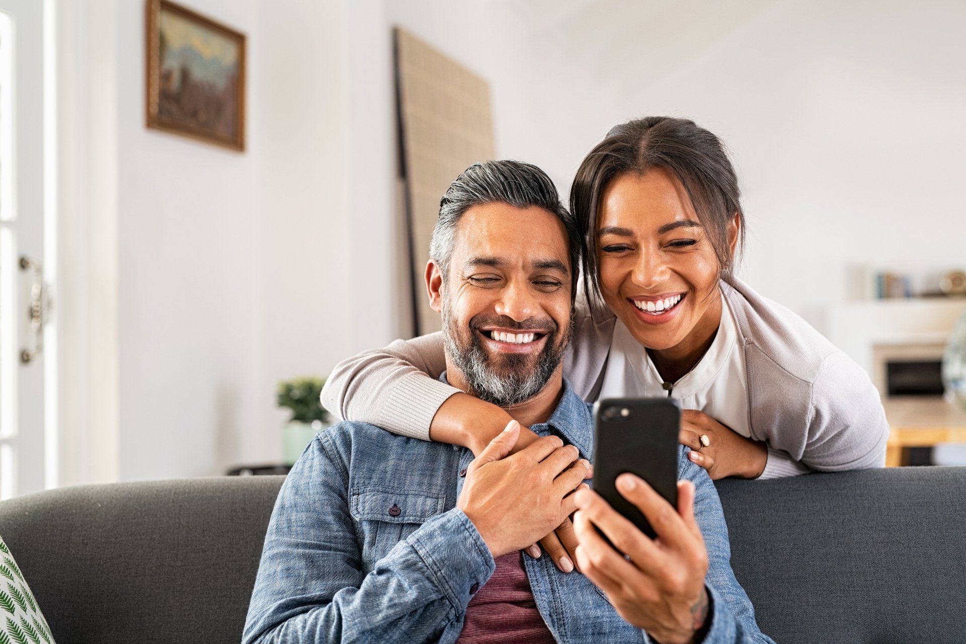 Two smiling people on a couch looking at a smartphone in a bright living room