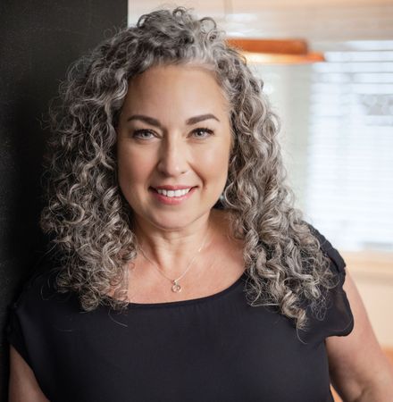 Smiling woman with curly gray hair in a black top, standing indoors near a window.