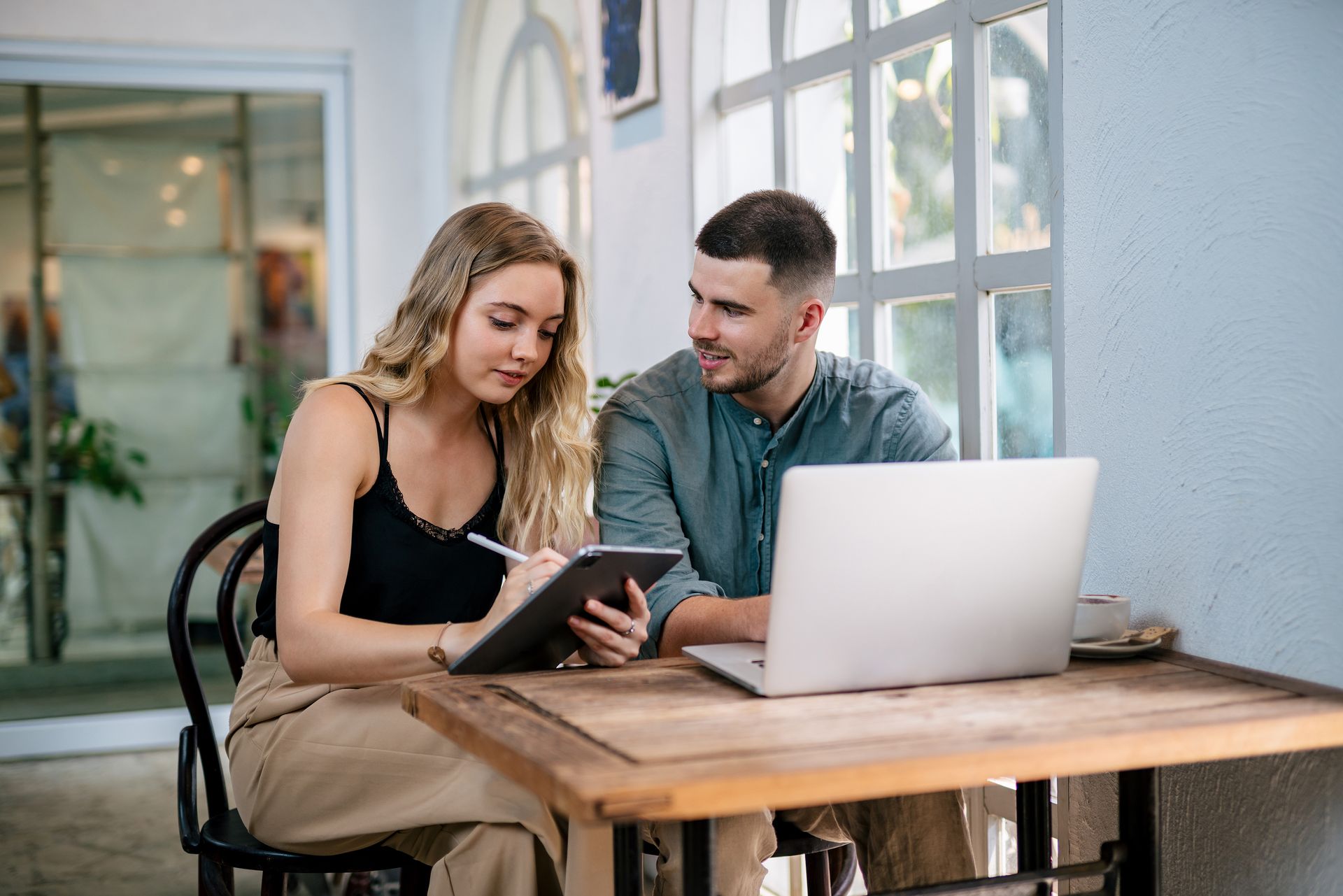 Two people reviewing a tablet at a cafe table with a laptop and coffee mugs