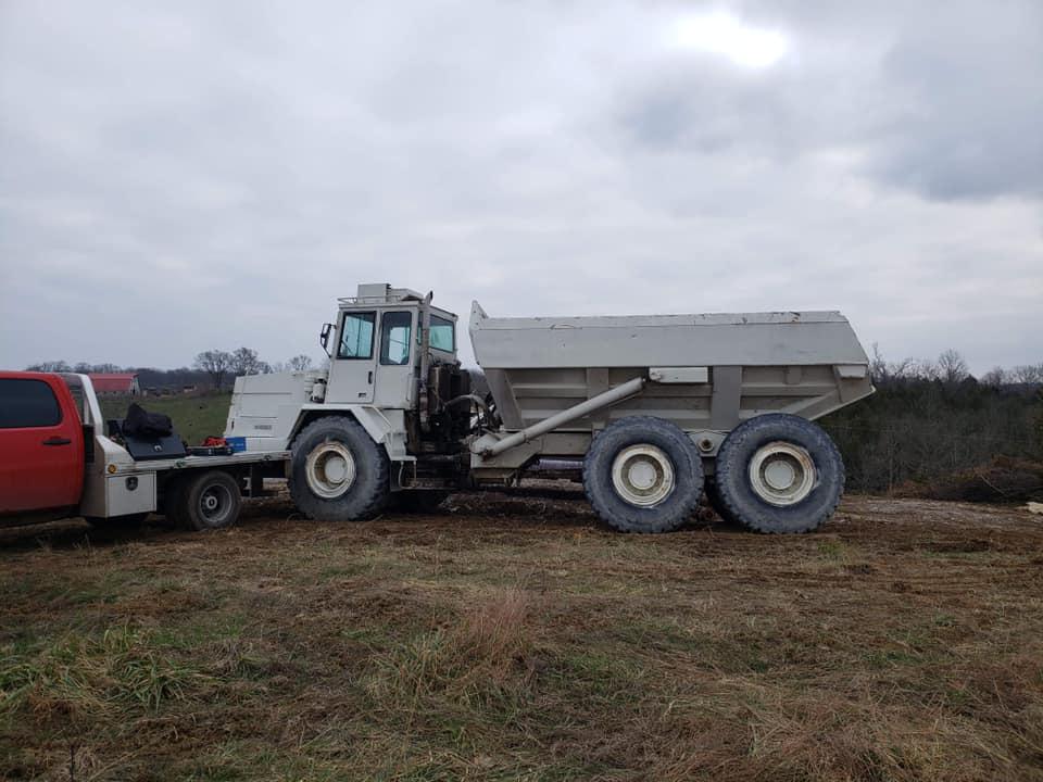 A dump truck is being towed by a red truck in a field.