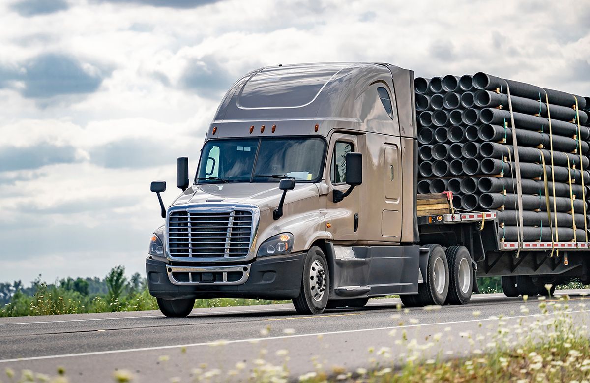 Semi-truck carrying black pipes on a highway, under a cloudy sky.