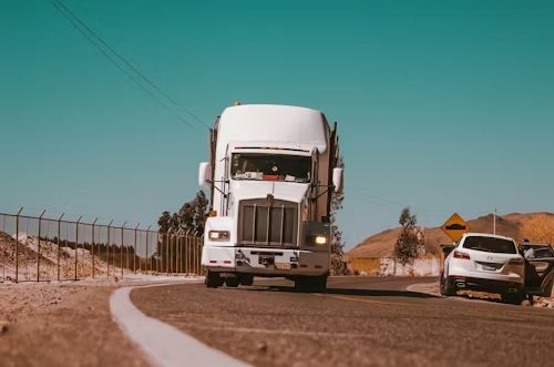 White semi-truck drives on a curved road, alongside a white car, against a blue sky.