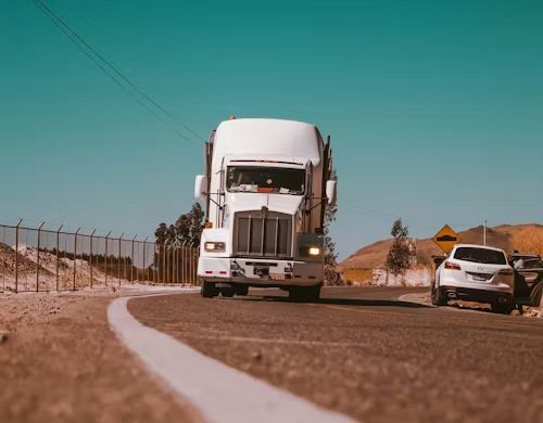 White semi-truck driving on a rural road beside a damaged car under a clear blue sky