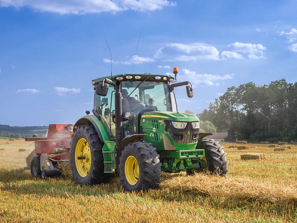 A green tractor is driving through a field of hay.