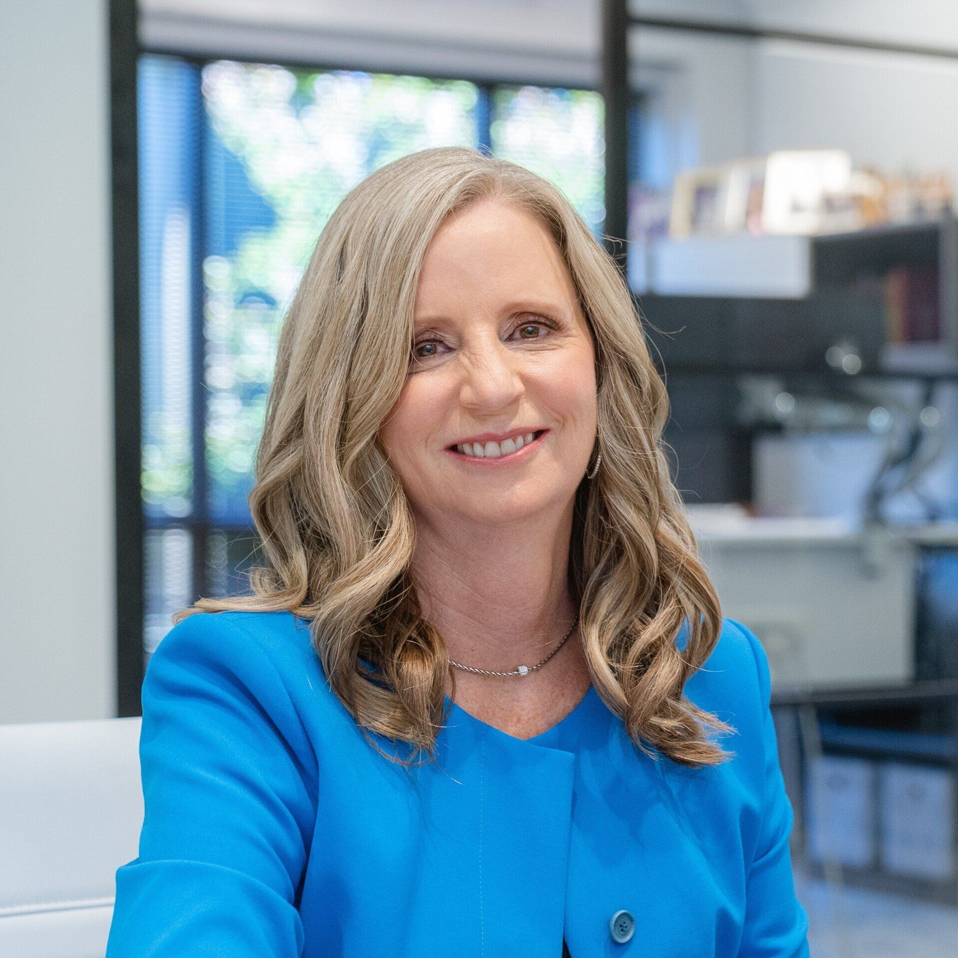 Woman with long, wavy blonde hair smiles, wearing a blue blazer, in an office setting.