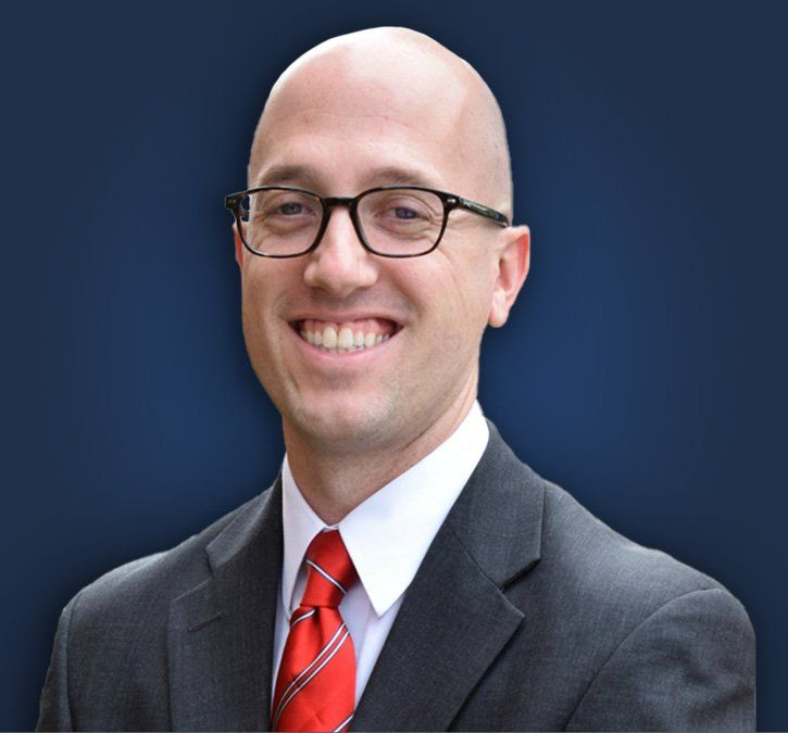 Man in glasses, suit, and red tie smiling, with a blue background.
