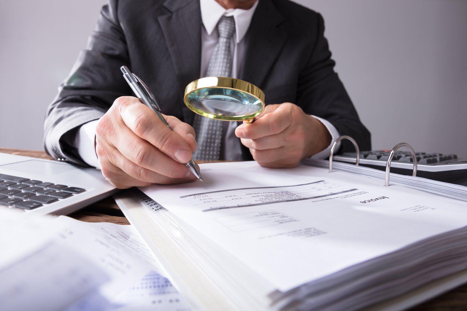 Person in suit using magnifying glass and pen to examine documents on desk.