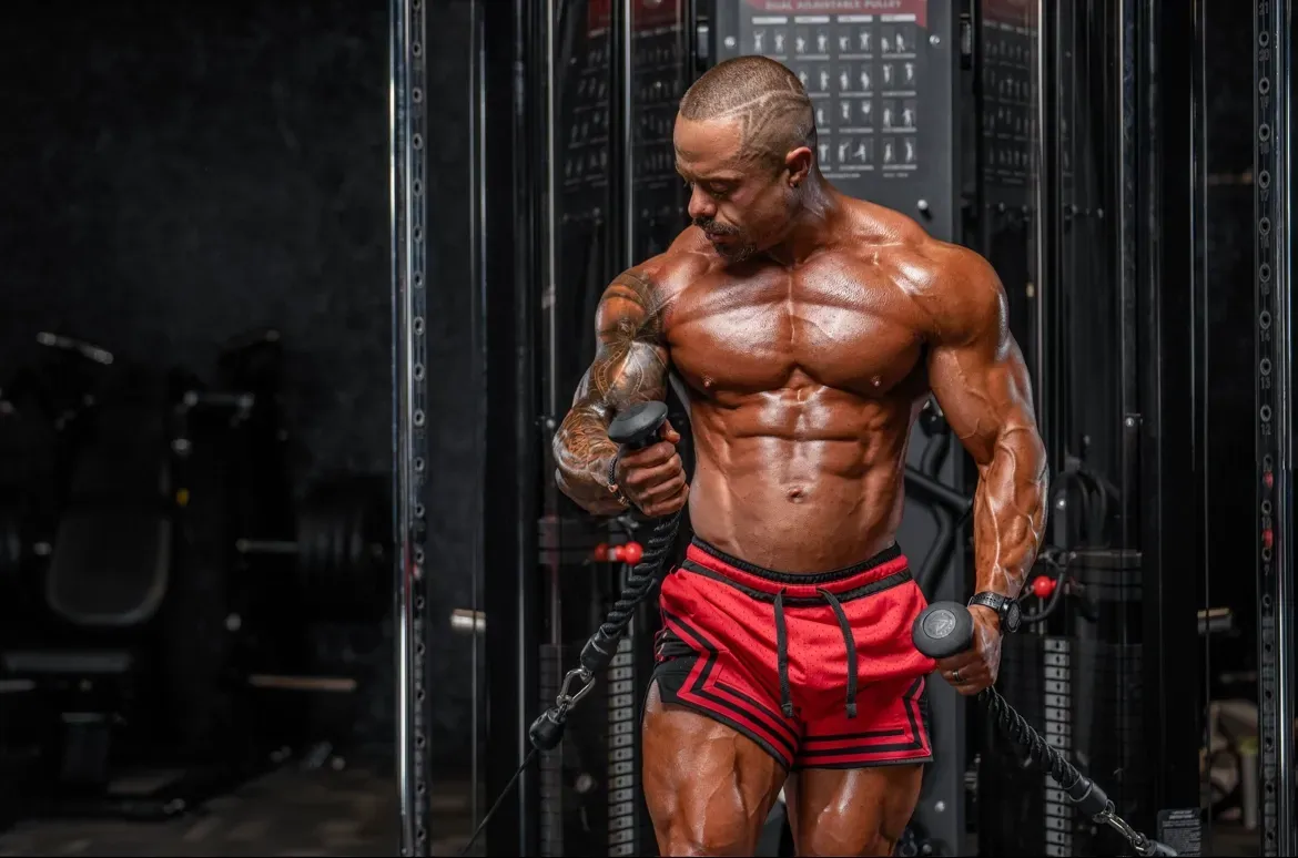 Muscular bodybuilder posing in a gym, wearing red shorts and holding dumbbells.