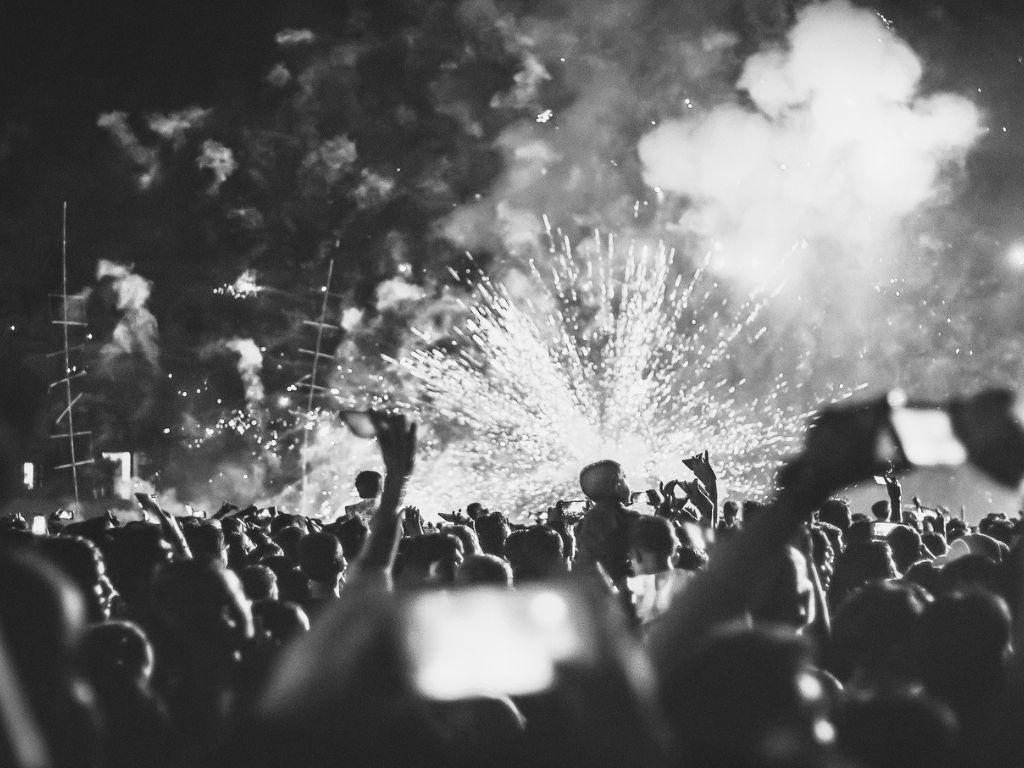 Foule lors d'un concert avec des feux d'artifice explosant au-dessus, les gens levant les bras et tenant des téléphones.