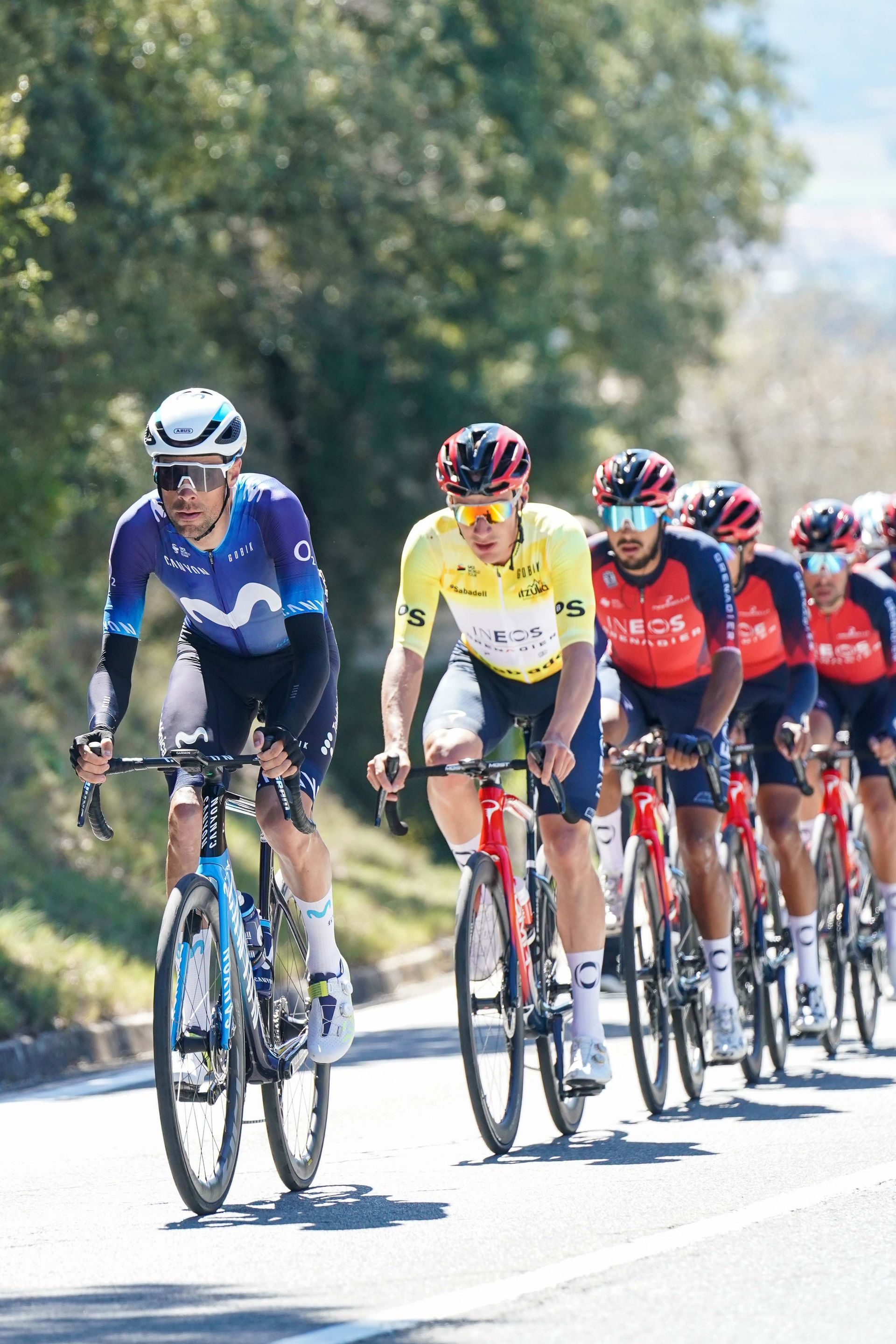 Des cyclistes courent sur une route ; l'un en jaune mène, suivi d'autres en maillot bleu, rouge et noir. Journée ensoleillée.