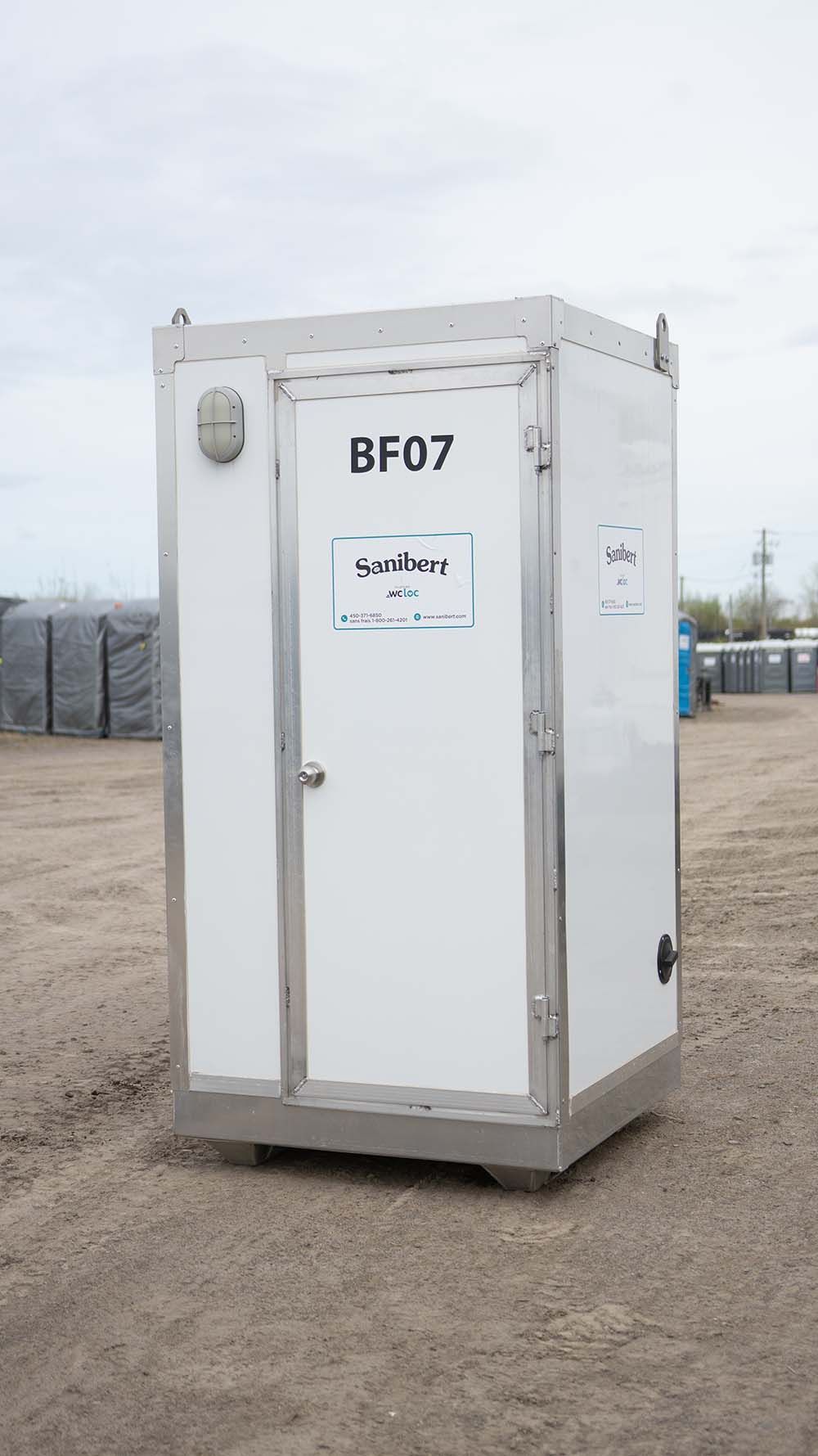 Toilettes portables blanches avec « BF07 » sur la porte, garées sur du gravier, ciel couvert.