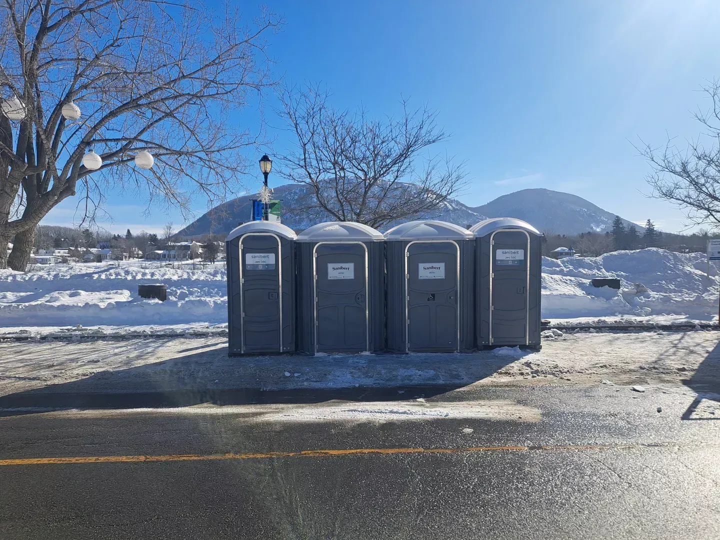 Quatre toilettes portables sont installées au bord d'une route enneigée, avec des montagnes et un ciel bleu azur en arrière-plan.