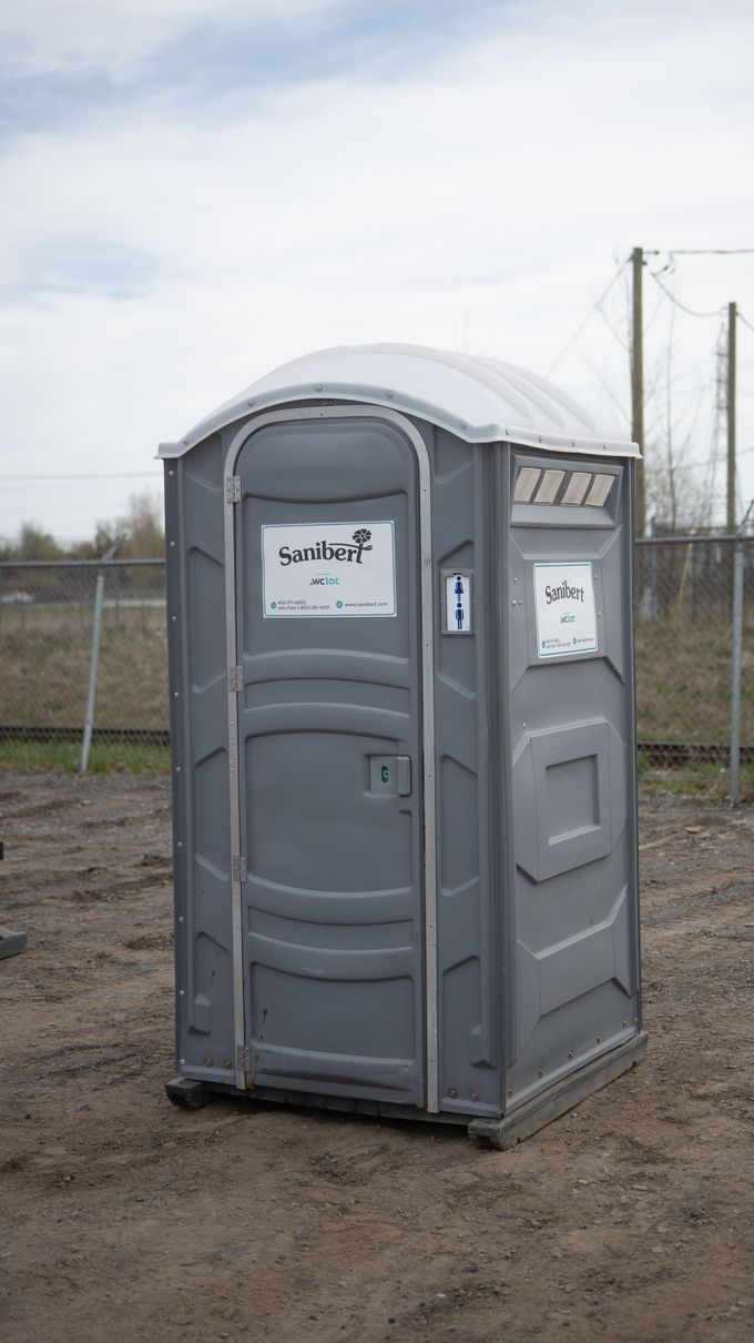 Toilettes portables grises sur sol en terre battue, à l'extérieur.