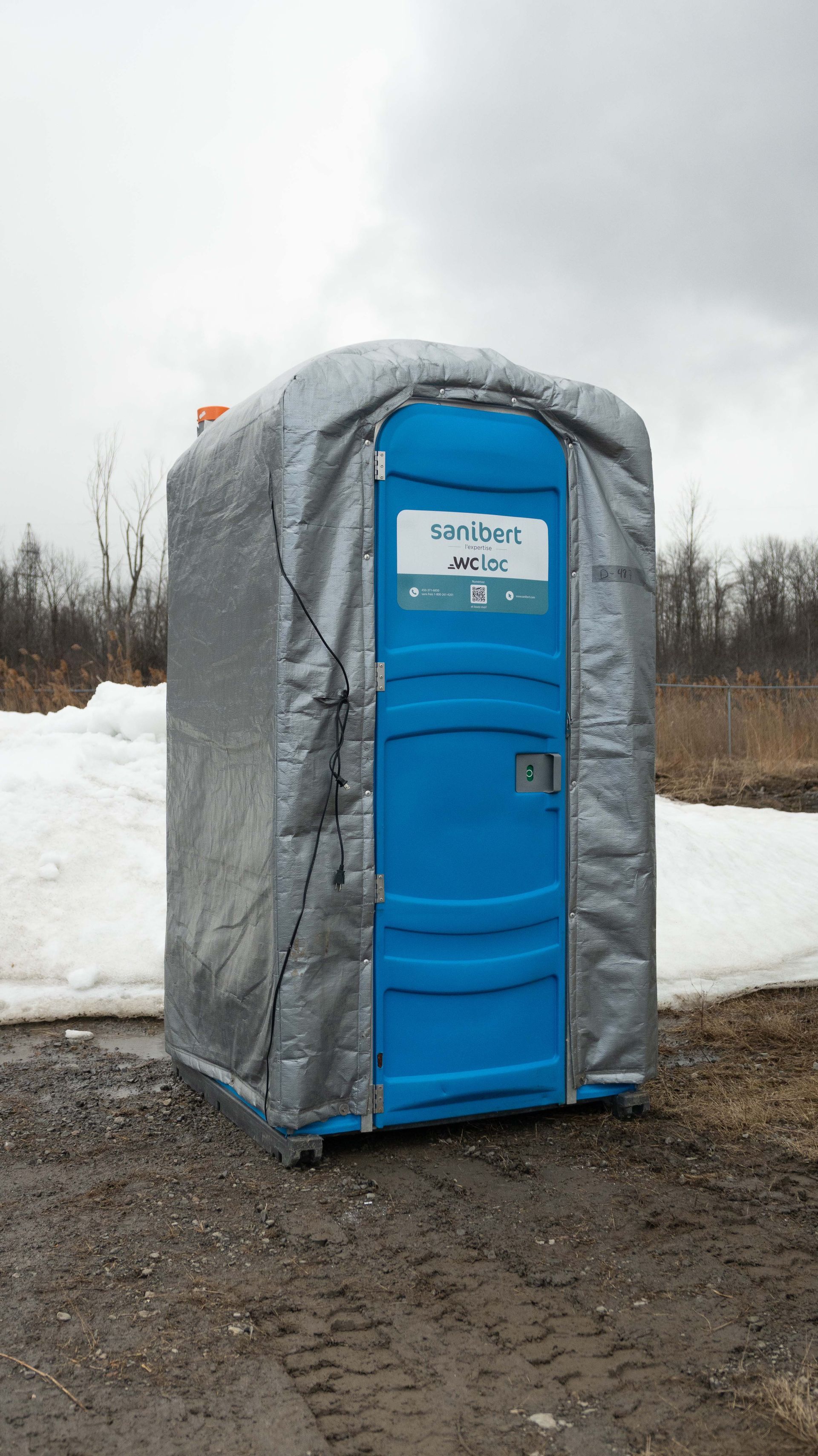 Blue portable toilet covered in silver insulation; snow and barren landscape in the background.