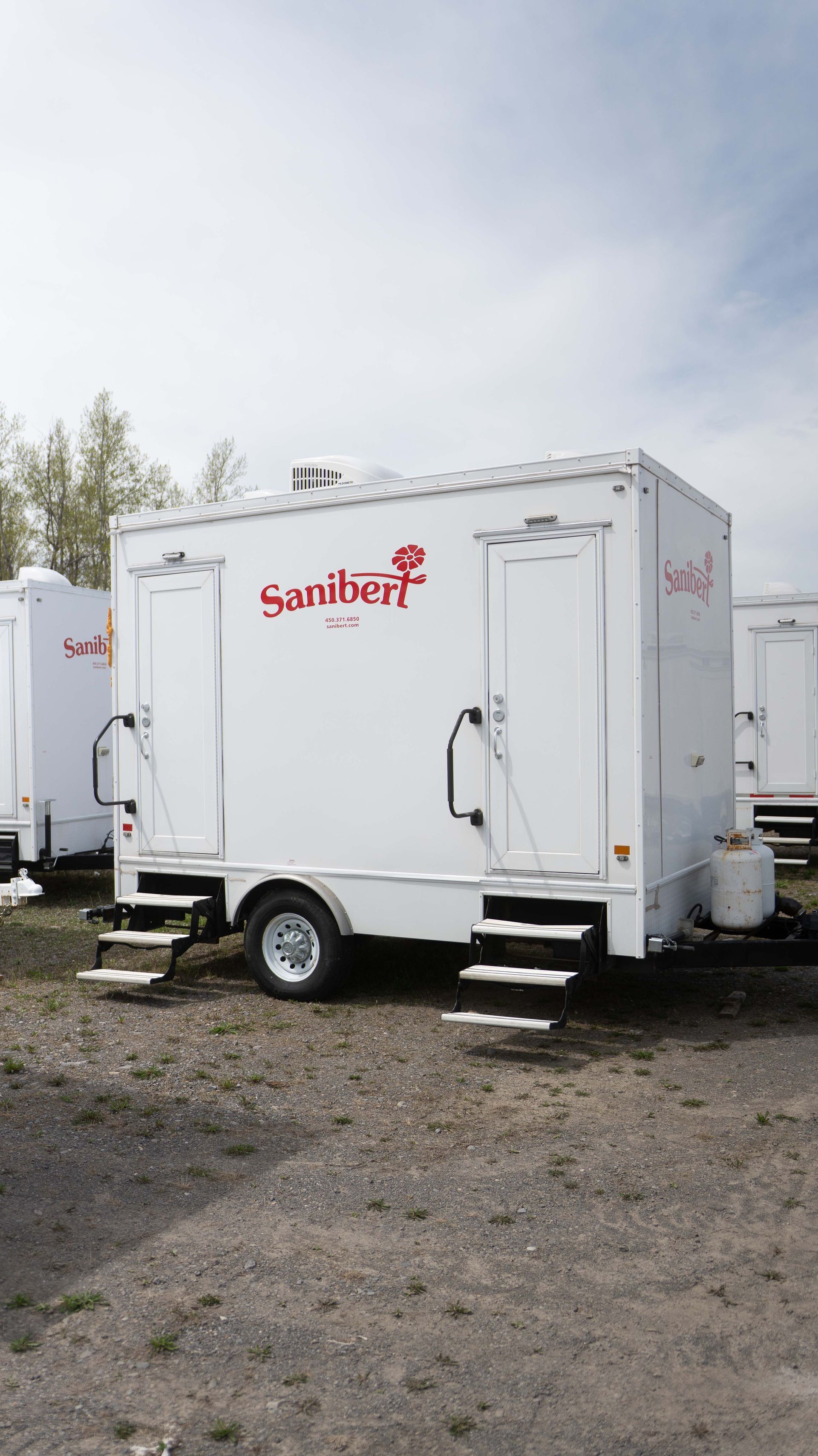 Toilettes mobiles blanches sur remorque. Comprend un marchepied et deux portes. Le logo Sunberg est visible sur le côté.