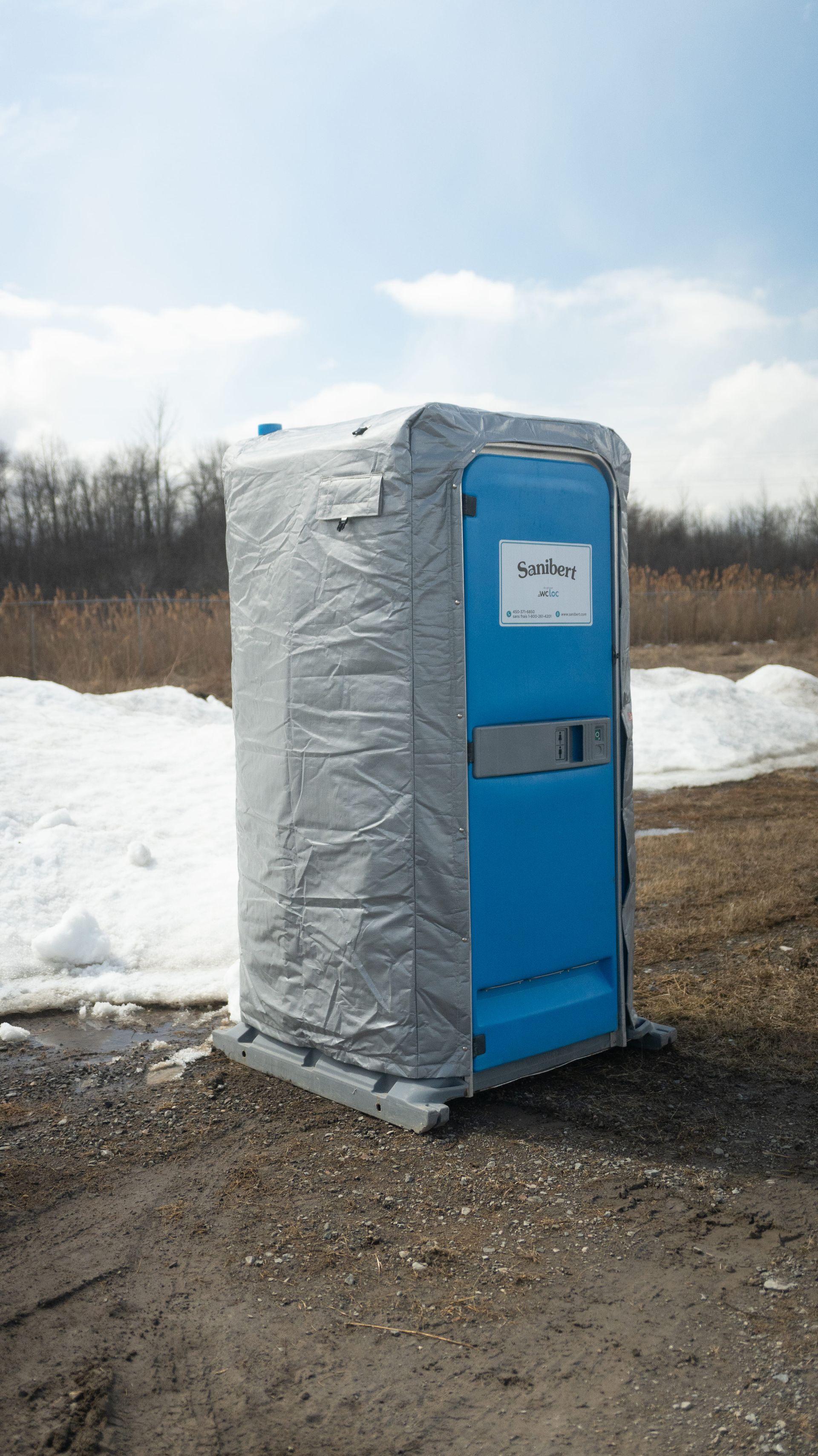 Blue portable toilet wrapped in gray plastic, on a gravel surface with snow and trees in the background.