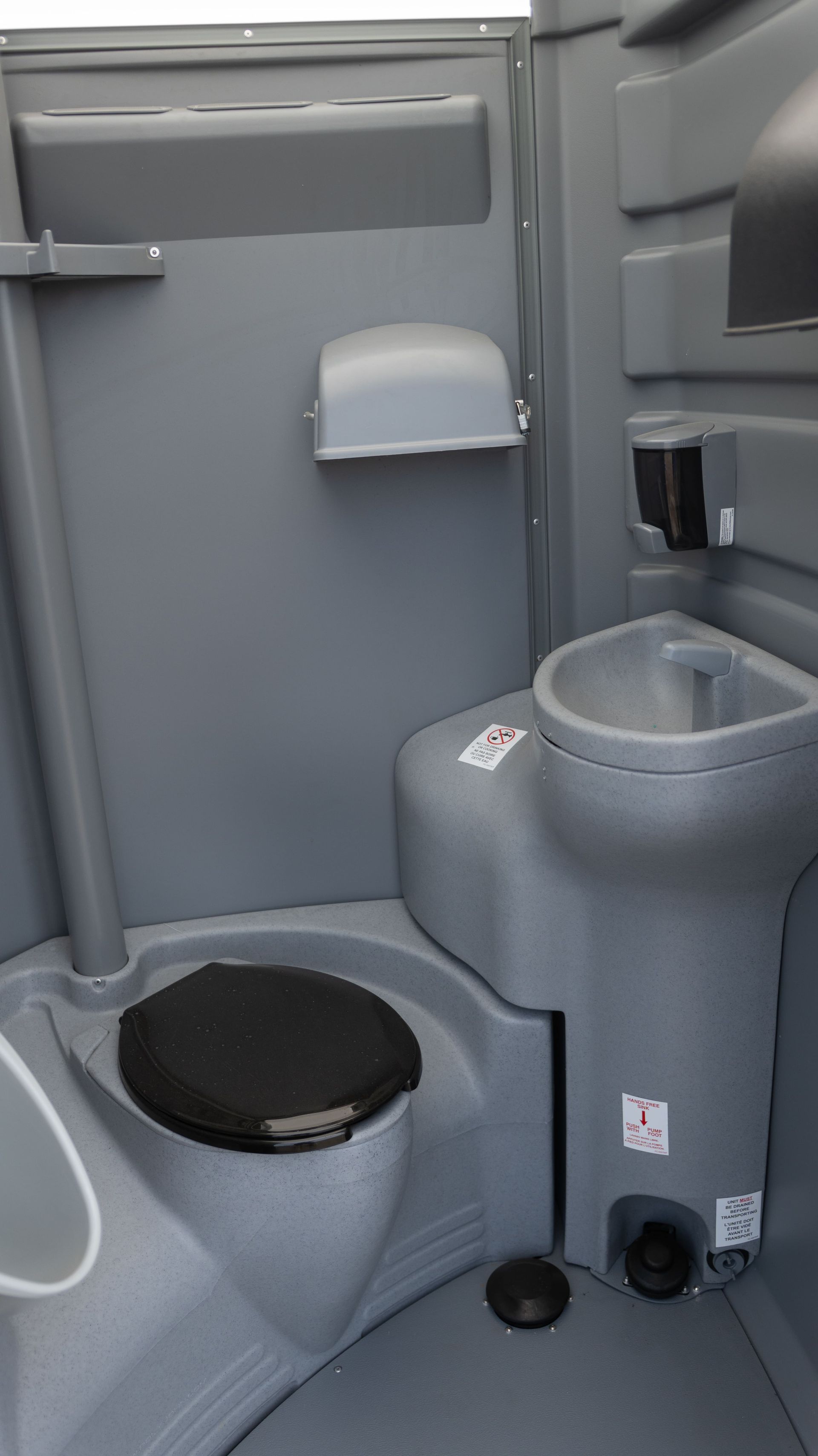 Des toilettes portables grises avec un panneau se trouvent à l'extérieur sur une surface en terre.