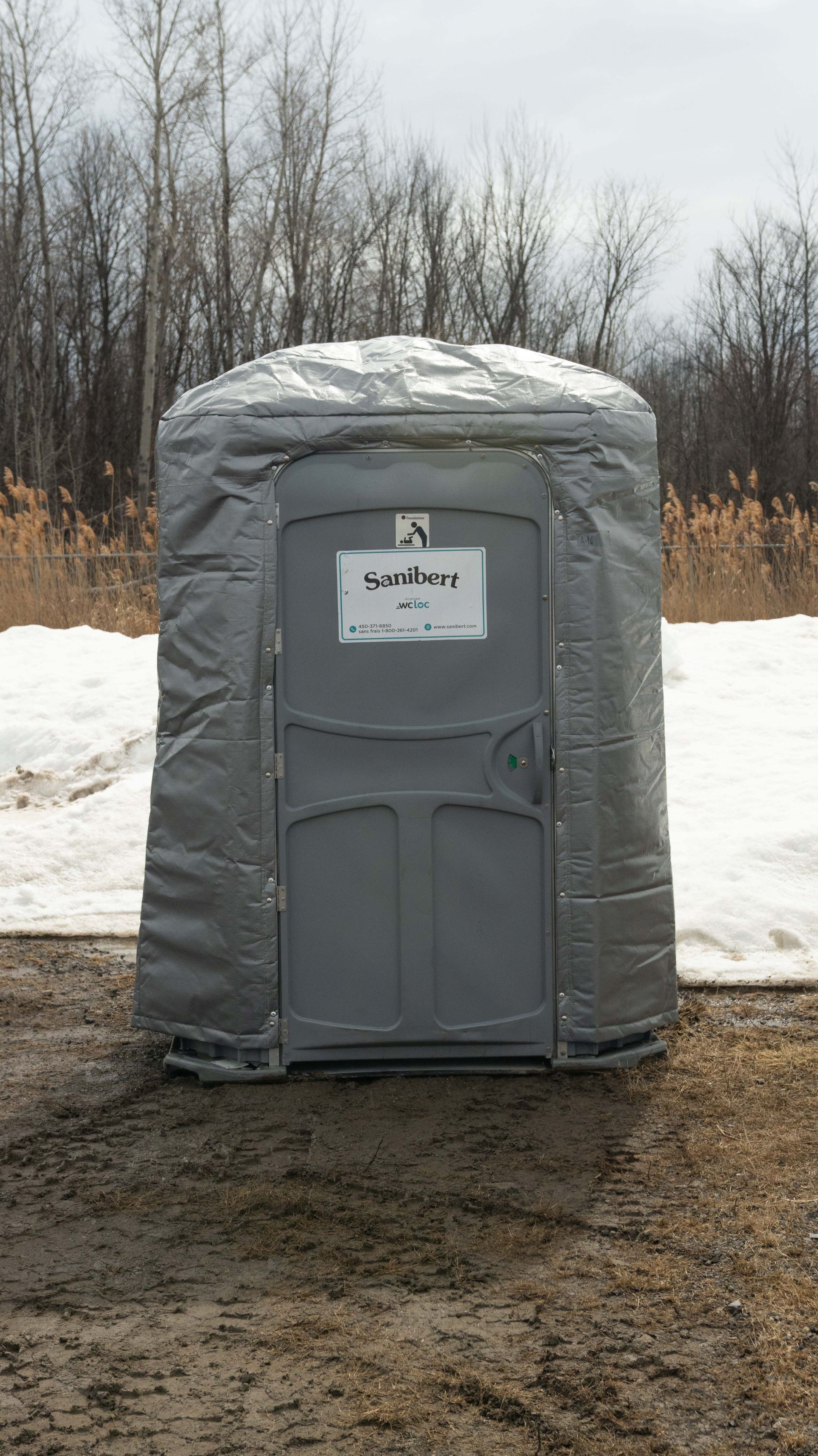 Gray portable toilet in a snowy, outdoor setting with a tarp wrapped around it.