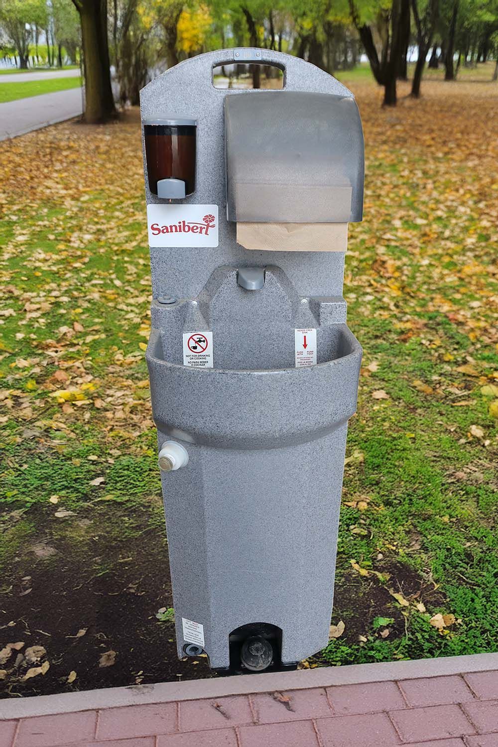Station de lavage des mains extérieure avec distributeur de savon, serviettes en papier et bouteilles d'eau, située dans un parc.