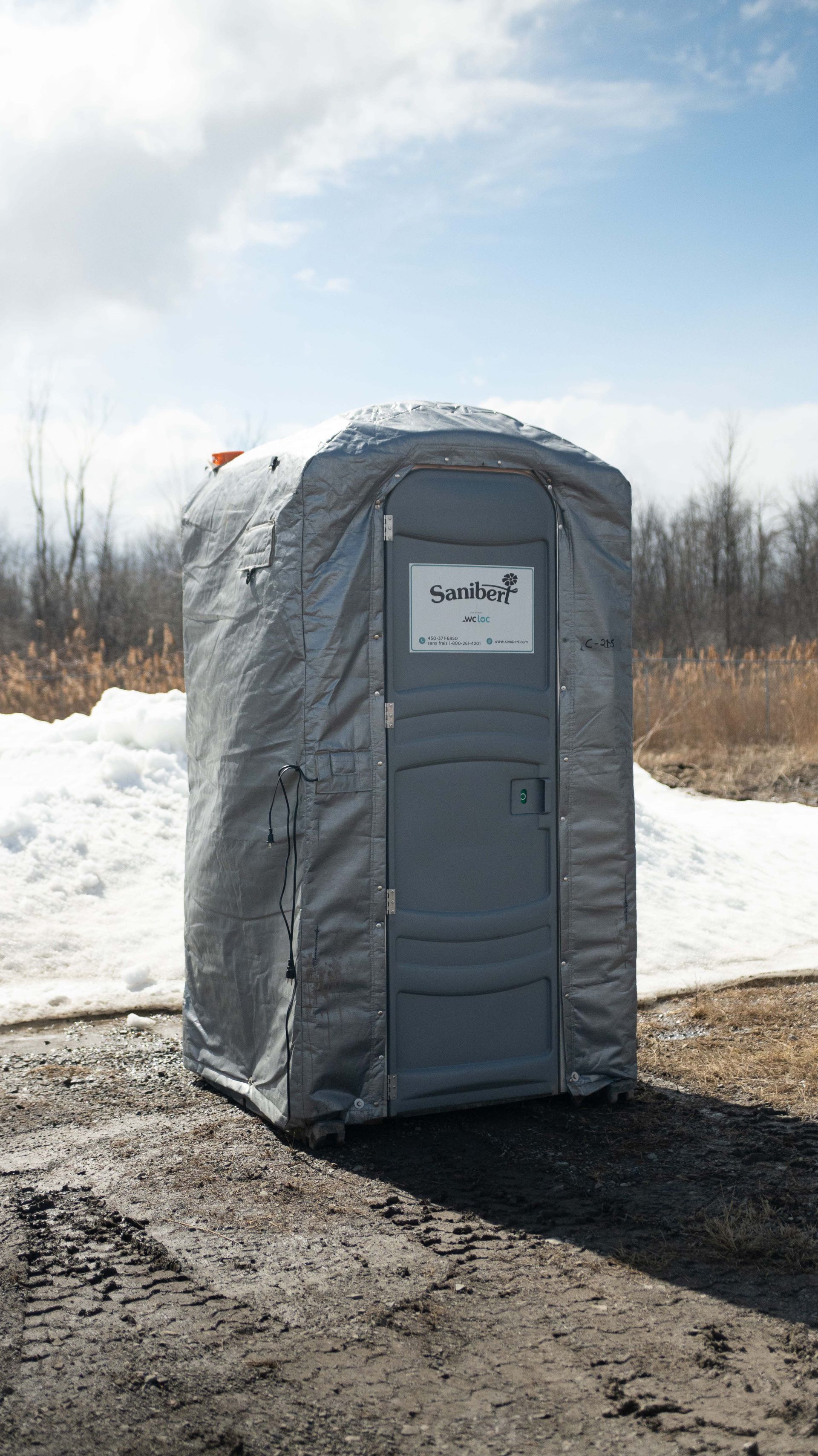 Gray portable toilet on a gravel surface, surrounded by snow and a sunny sky.