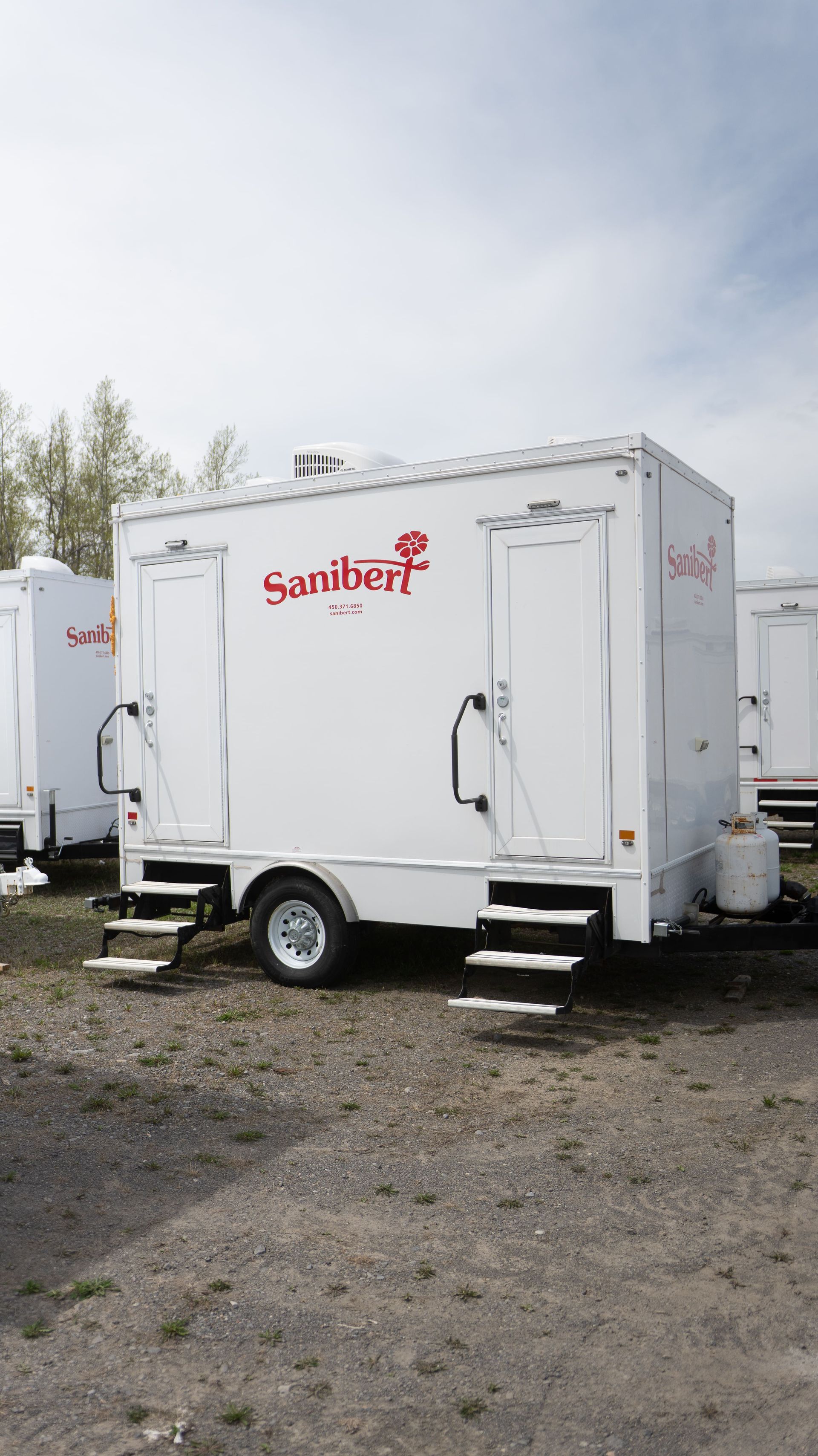 White portable restroom trailer with Sanibon logo. Two entrances with steps. Gravel lot, cloudy sky.