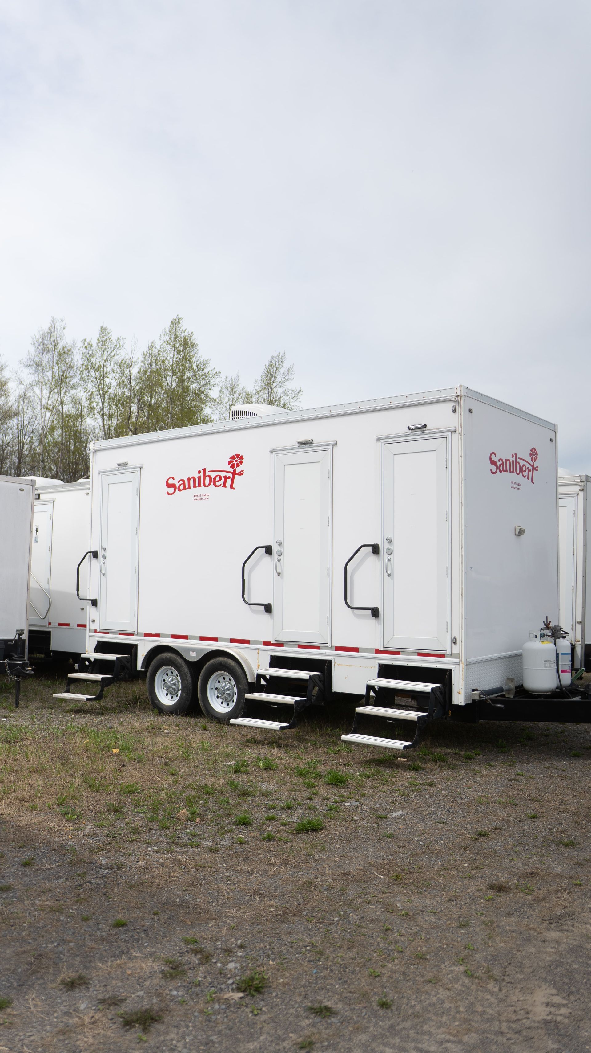 Remorque de toilettes portables blanches avec marches, logos rouges, stationnée à l'extérieur.
