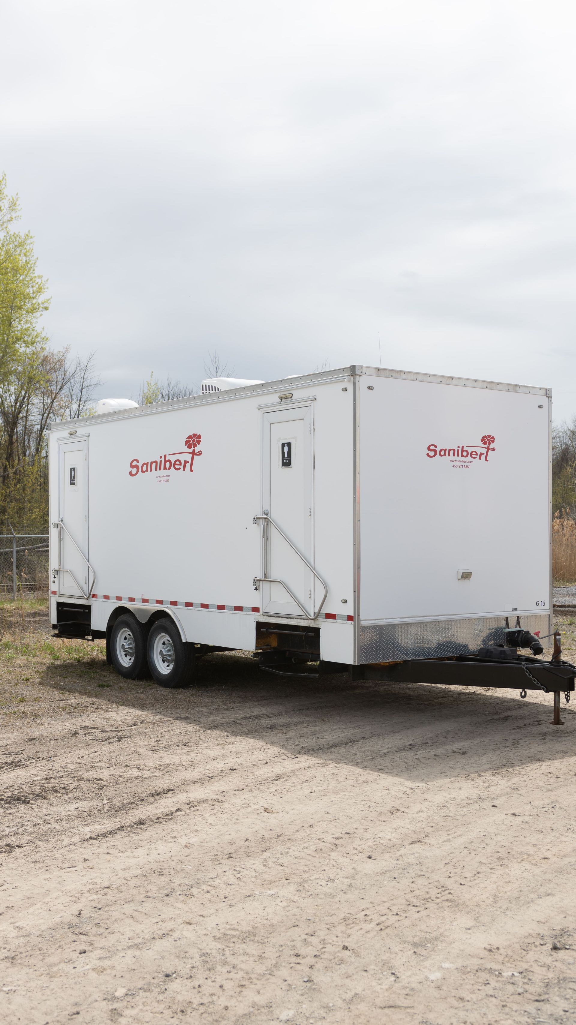 White portable restroom trailer on a gravel lot; red logo on the side.