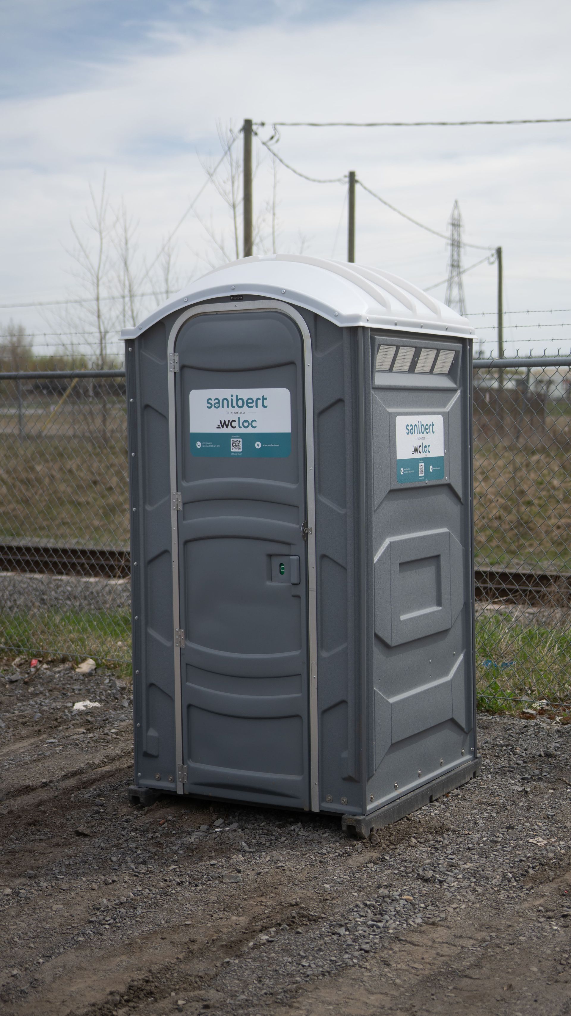 Toilettes portables grises dans une zone de gravier avec une clôture, des lignes électriques et un ciel nuageux en arrière-plan.