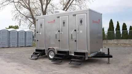 Toilettes mobiles sur un terrain en gravier. Avec plusieurs cabines, un revêtement argenté et des marches. Arbres en arrière-plan.