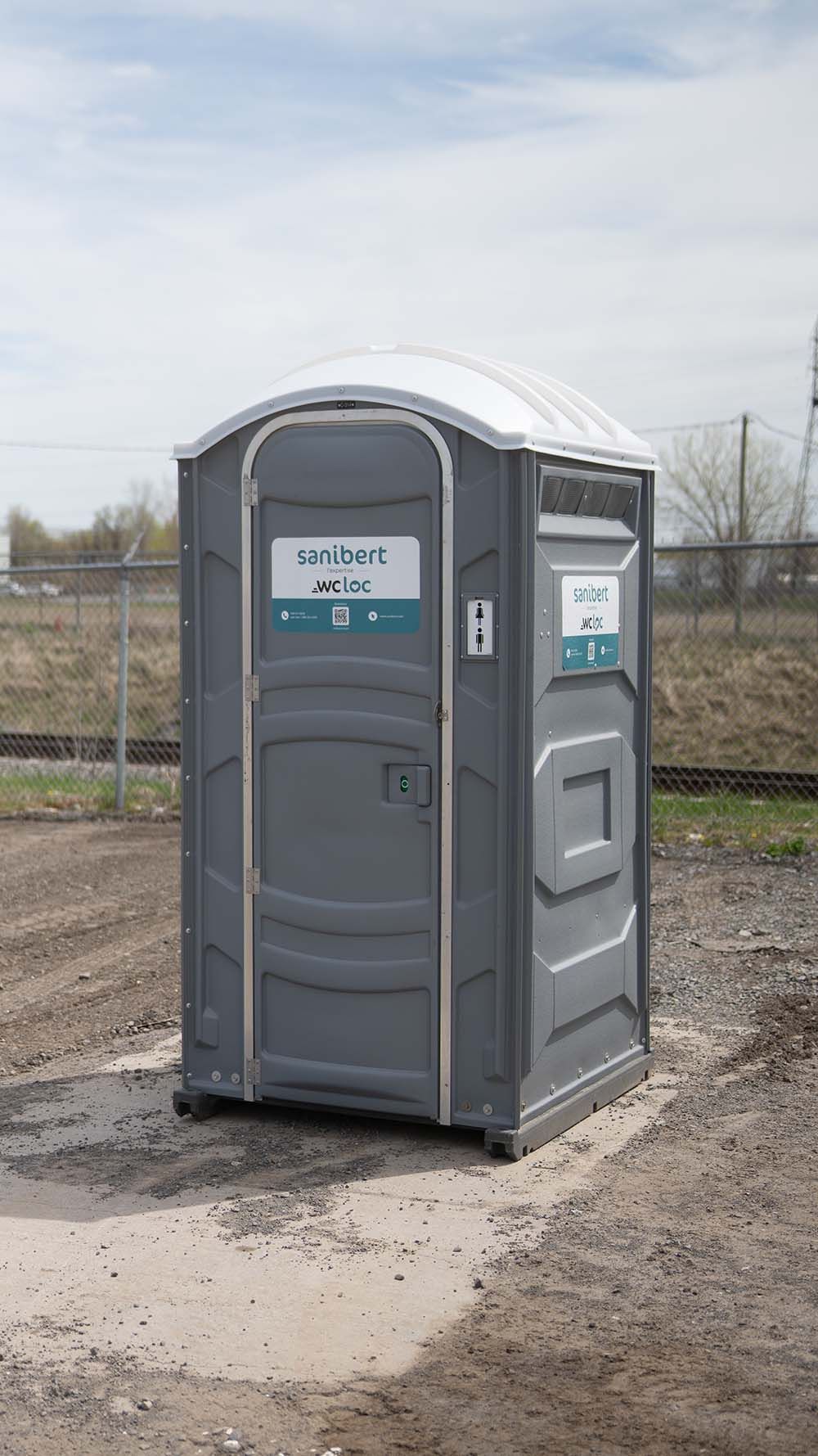 Toilettes portables grises sur un chemin de terre, situées à l'extérieur.