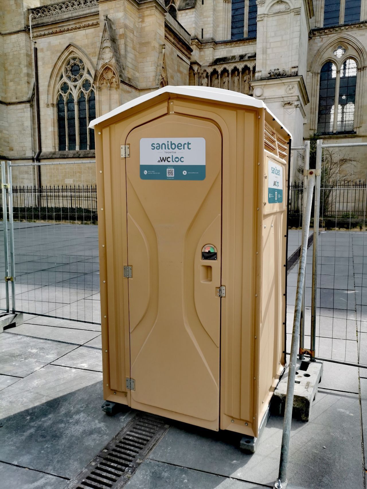 Toilettes portables beiges devant une cathédrale, à côté d'une balustrade en métal.