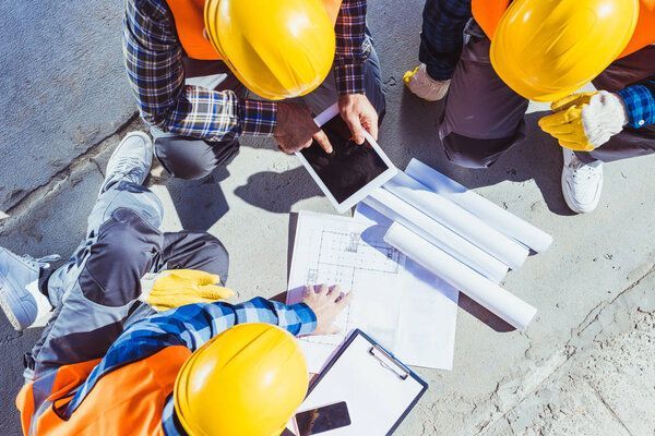 Des ouvriers du bâtiment portant des casques et des gilets de sécurité examinent des plans et une tablette à l'extérieur.