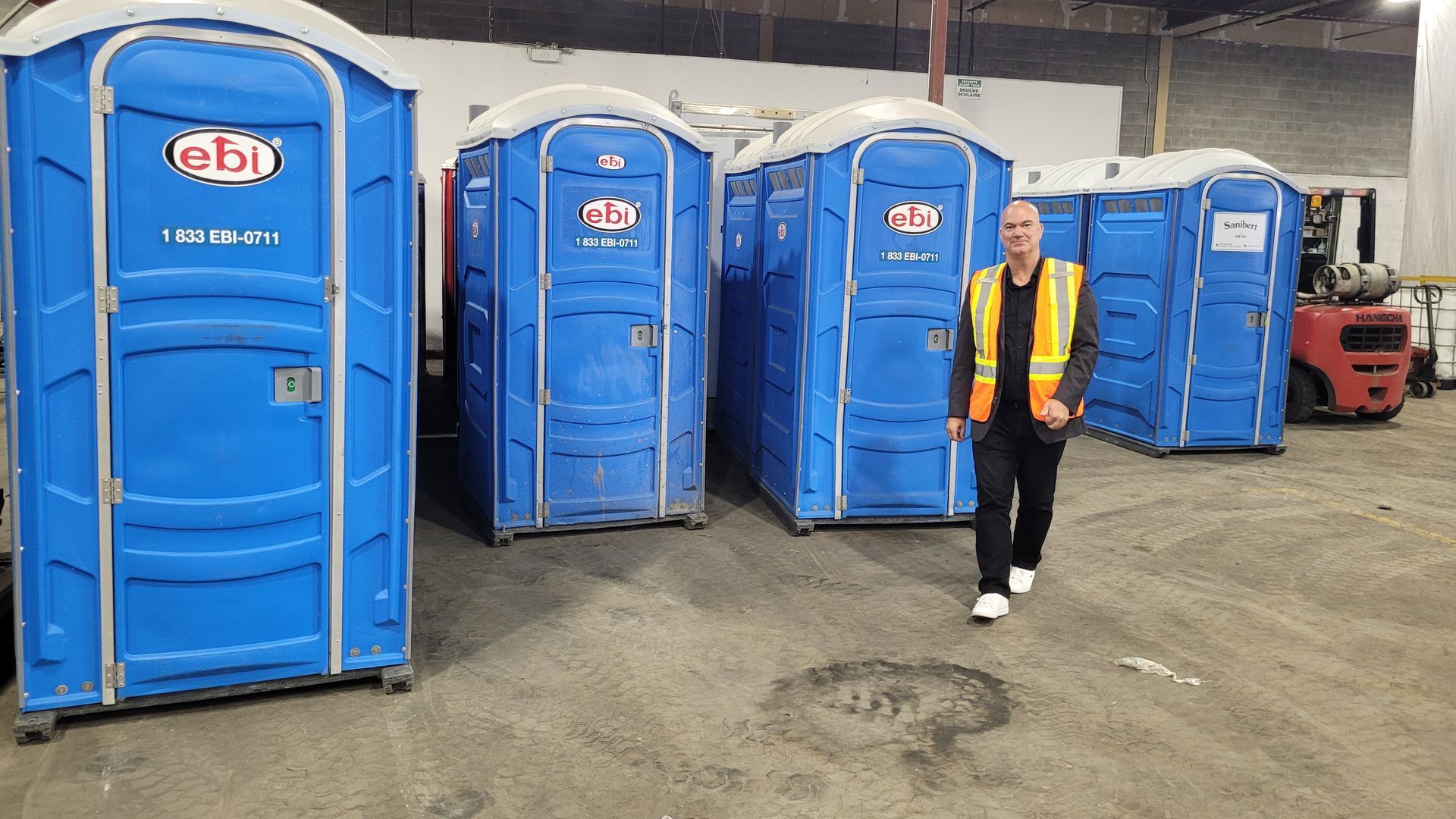 Un homme passe devant des toilettes portables bleues dans un entrepôt. Il porte un gilet de sécurité.