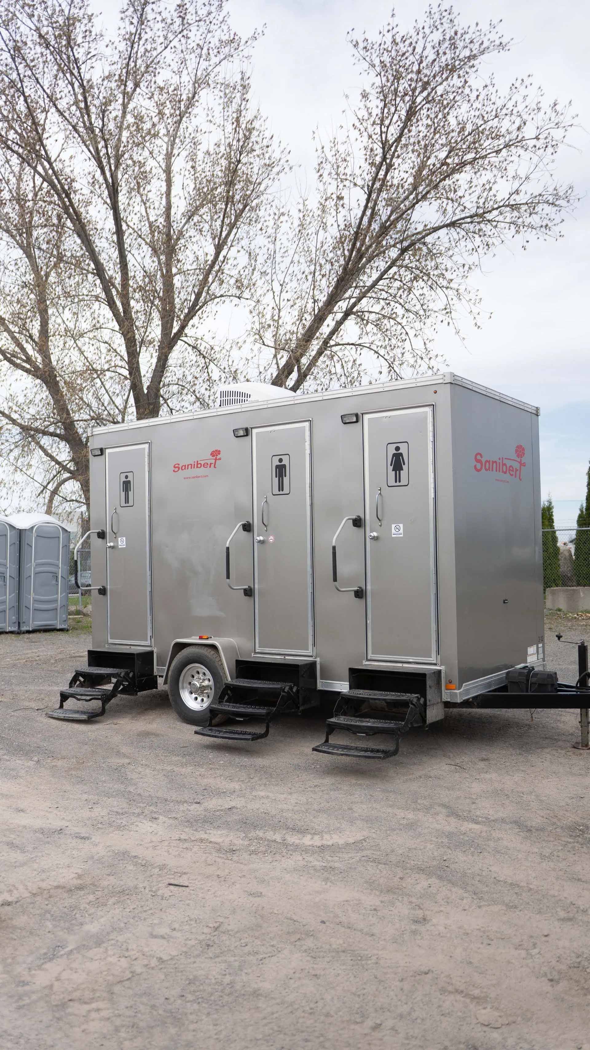 Silver portable restroom trailer with three individual stalls on a trailer in an outdoor setting.