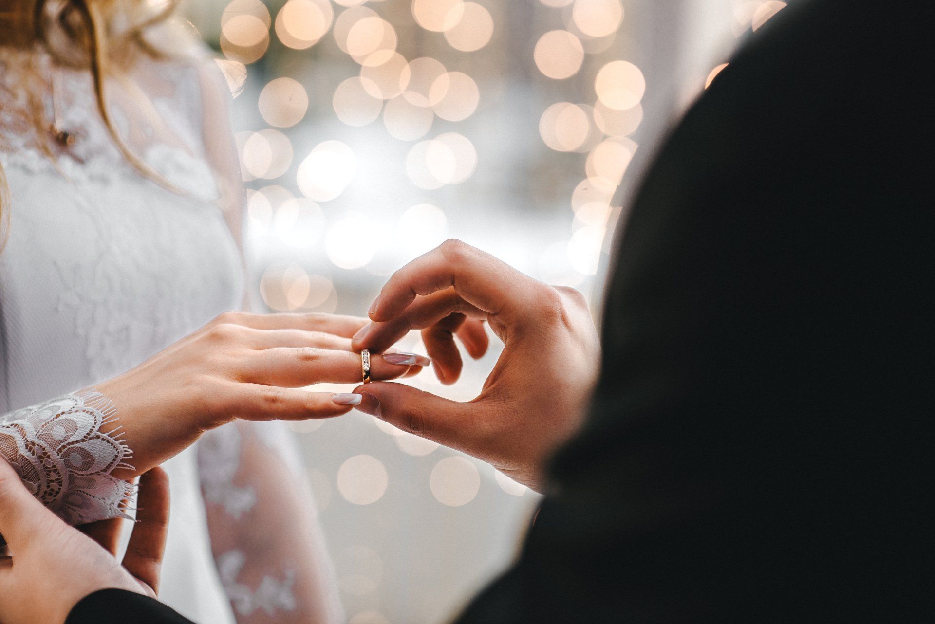 Un homme place une alliance sur le doigt d'une femme ; tous deux sont habillés pour un mariage, avec des lumières floues en arrière-plan.