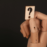 A wooden mannequin hand holding a small, light-colored square sign with a black question mark against a black background.