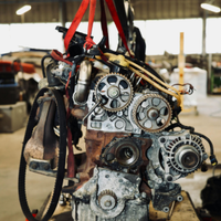 A car engine hanging from a crane in an industrial workshop, held by red straps.