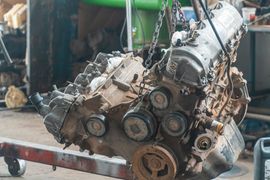 An automotive engine hanging from a chain hoist in a cluttered workshop.