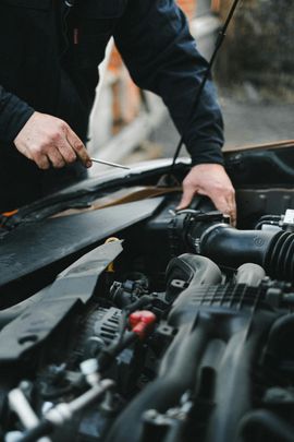 A person in a dark jacket works on the engine of a vehicle, holding a tool while reaching into the open hood.