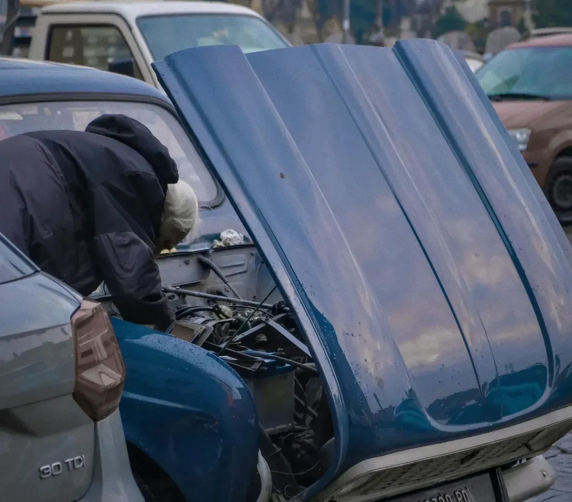 A person in a dark jacket leans under the open hood of a blue vintage car parked on a wet city street.