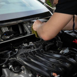 A mechanic cleans the area beneath a car's windshield with a yellow cloth while working on the engine.