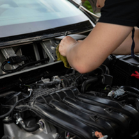 A mechanic cleans the area beneath a car's windshield with a yellow cloth while working on the engine.