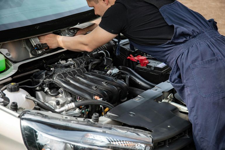 A mechanic in dark blue coveralls working under the hood of a car, reaching toward the back of the engine bay.