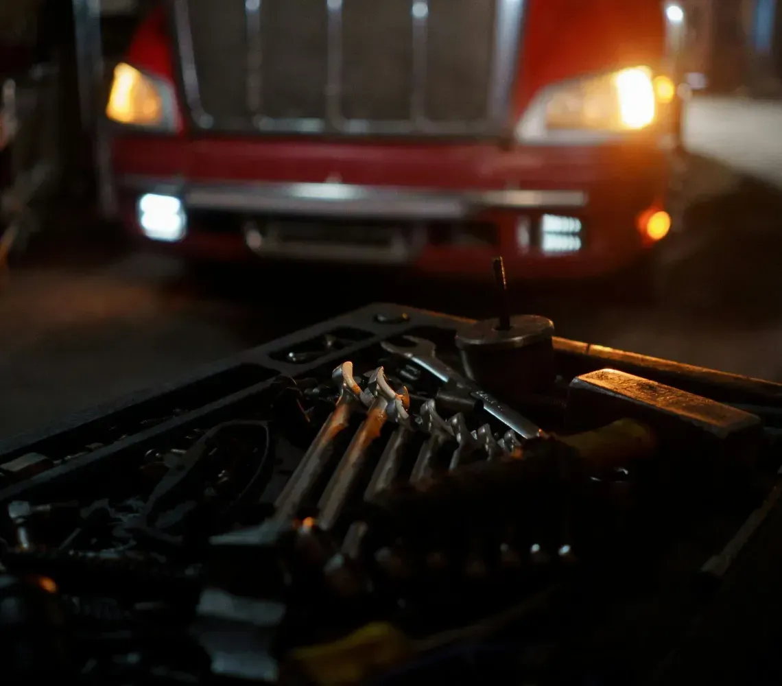 A red semi-truck stands in a dark garage behind a tool tray containing assorted wrenches and tools in the foreground.