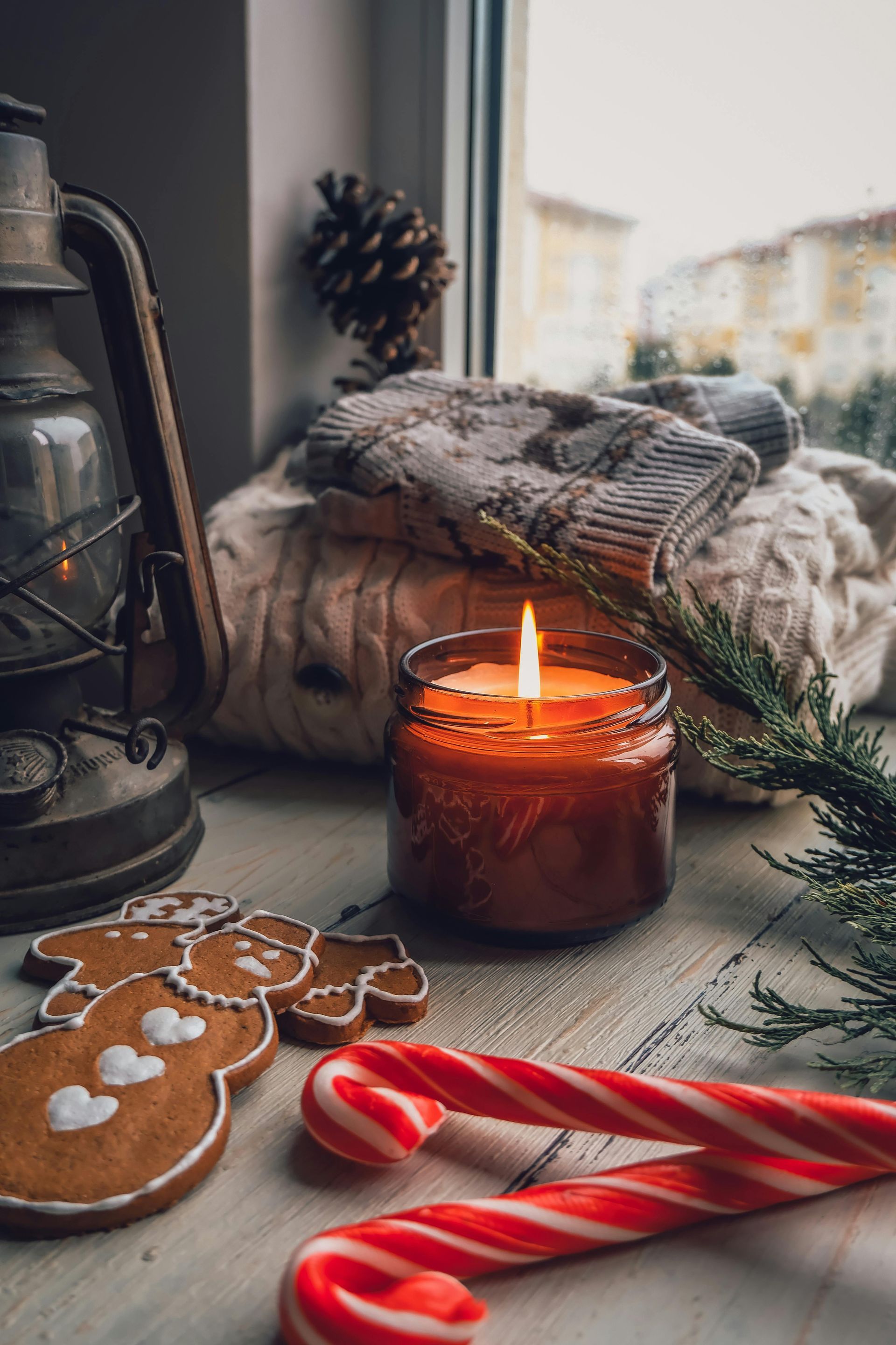 Candle burning near sweaters, gingerbread cookies, candy canes, and a lantern on a windowsill.